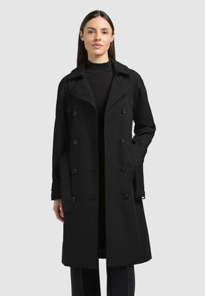 Kvinne iført en lang, svart dobbeltspent trenchcoat med belte, stående mot en ensfarget lys grå bakgrunn.