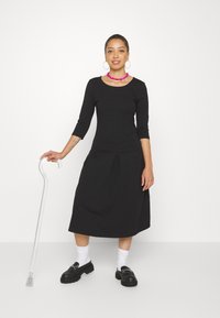 Musta, vartalonmyötäinen mekko, jossa on kolme neljäsosaa hihat ja laskostettu hame. Yhdistetty mustiin kenkiin, valkoisiin sukkiin ja selkeään keppiin. Vaaleanpunainen kaulakoru.