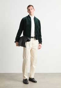 Chaqueta bomber de felpa verde sobre una camisa blanca de botones, pantalones de crema y zapatos de cuero negros, sosteniendo un bolso negro texturizado.