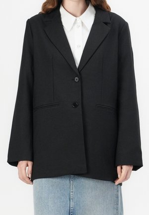 Frau trägt einen schwarzen maßgeschneiderten Blazer über einem weißen, geknöpften Hemd und einem hellblauen Jeansrock, von Hals bis Hüfte gezeigt.