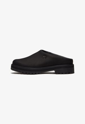Scarpa mule nera slip-on con suola spessa e scolpita e piccolo logo a forma di albero sul lato, mostrata dal lato esterno su sfondo bianco.