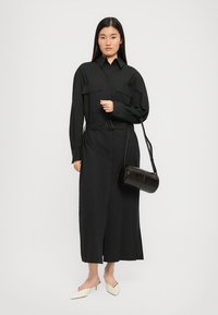 Black long-sleeve shirt dress with two chest pockets, belted waist, and side slit. Paired with a cylindrical black bag and white pointed shoes.