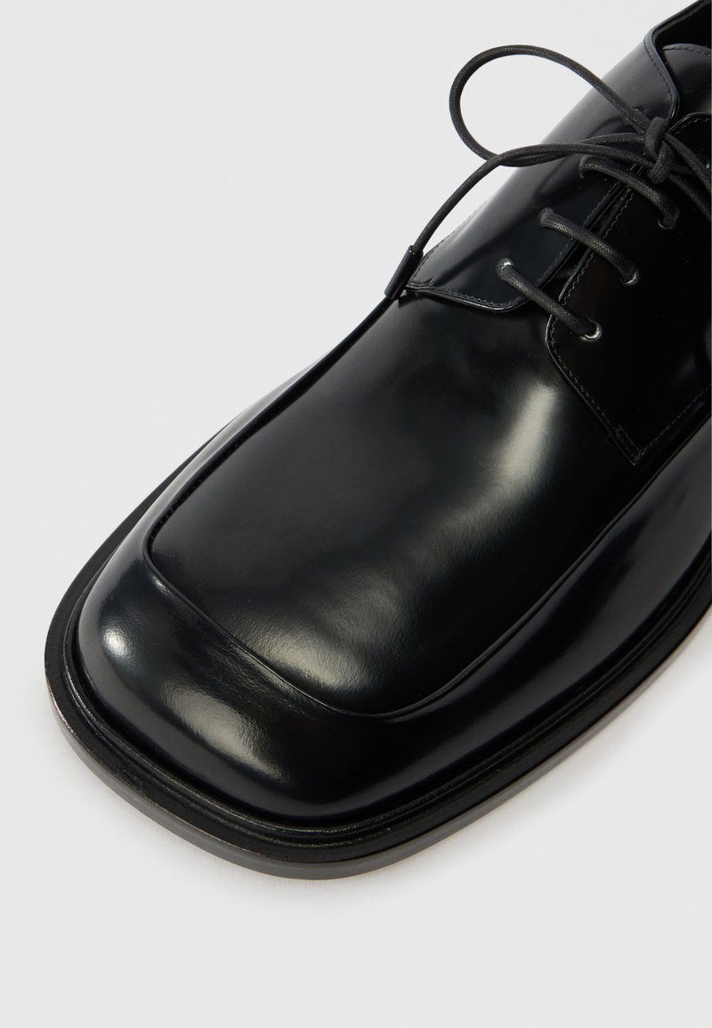 Scarpa di pelle nera con finitura liscia, punta arrotondata, cuciture in rilievo e lacci neri. La suola è leggermente elevata e arrotondata.