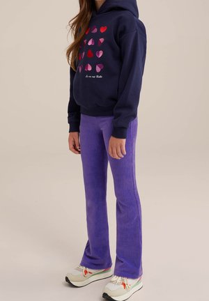 Young person wearing navy hoodie with multicolored hearts and French text, purple ribbed pants, and white sneakers with colored accents.