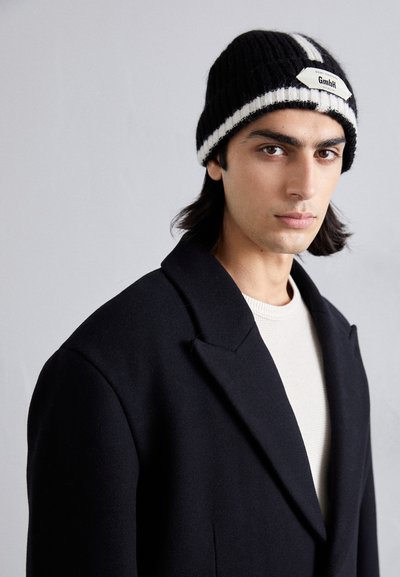 Bonnet tricoté à rayures noires et blanches avec un patch logo, porté avec un manteau noir et une chemise texturée de couleur claire. Tissu doux et coupe décontractée.