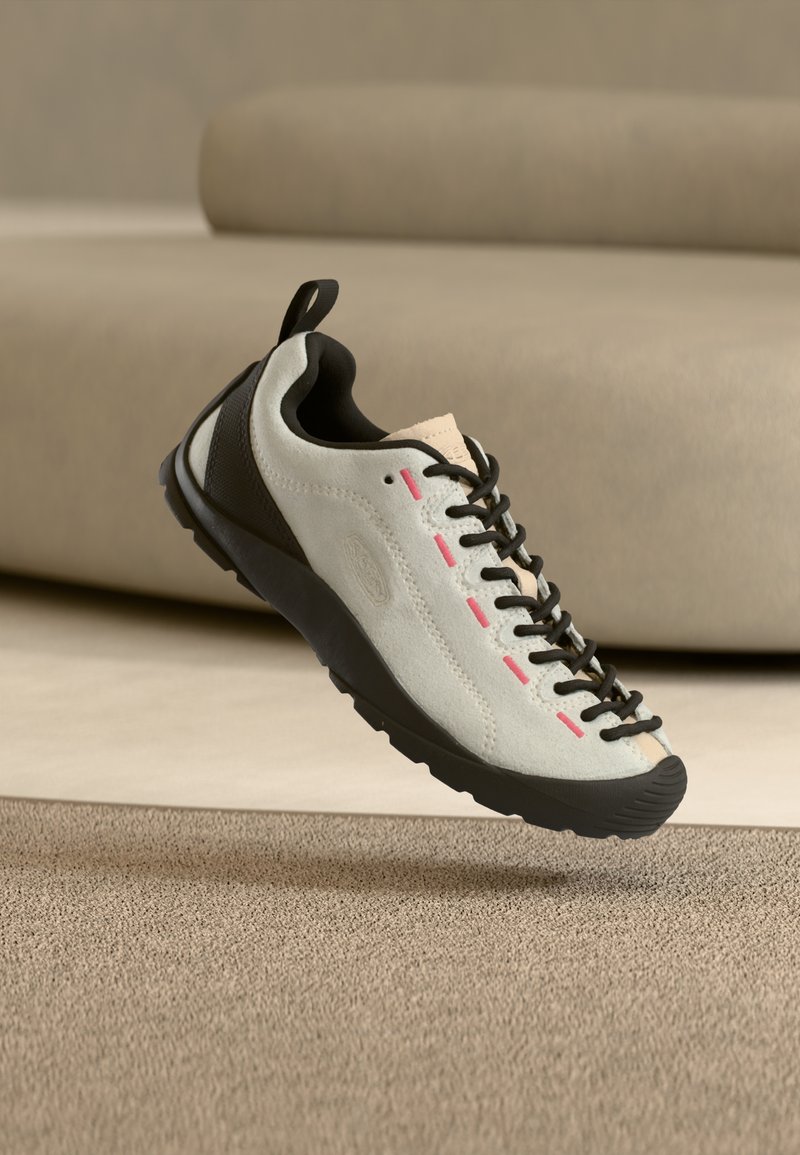 Λευκό και μαύρο αθλητικό παπούτσι με ροζ ραφές, μαύρα κορδόνια και υφή μαύρης σόλας να βρίσκεται πάνω σε μπεζ χαλί με φόντο μπεζ καναπέ.