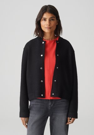 Black textured cropped jacket with silver snap buttons, worn over a red top, paired with gray jeans. Simple design, relaxed fit.