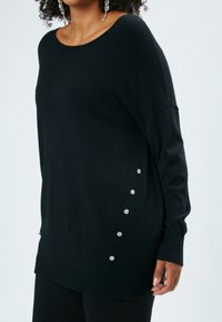 Maglione nero, comodo e largo, realizzato in un tessuto morbido. Presenta un ampio scollo, maniche lunghe e bottoni decorativi floreali in tono argento sul lato.