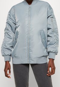 Giacca bomber azzurro chiaro realizzata in tessuto liscio e lucido, con polsini e orlo a costine, tasche laterali con zip e dettaglio del logo sul petto.