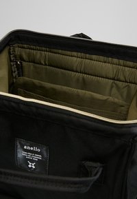 Sac en toile noire avec fermeture à glissière, intérieur vert olive et plusieurs compartiments. Étiquette portant le nom "anello" avec détails de couture.