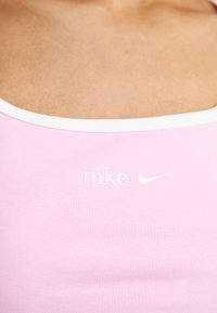 Růžový sportovní top s bílým lemem, s vyraženým logem "nike" a tikem. Materiál vypadá hladce a pružně, ukazuje jemnou texturu žebrování.