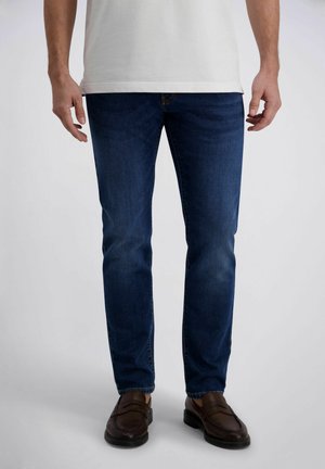 Mann trägt dunkelblaue Slim-Fit-Jeans, weißes Hemd und dunkelbraune Leder-Loafer, steht vor einem schlichten Hintergrund.