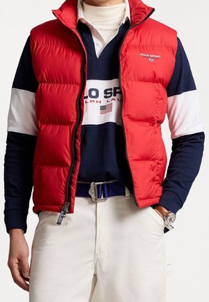 Man draagt een rode gewatteerde bodywarmer over een marineblauw-wit Polo Sport rugbyshirt met witte broek en blauwe riem, handen gedeeltelijk in de zakken.