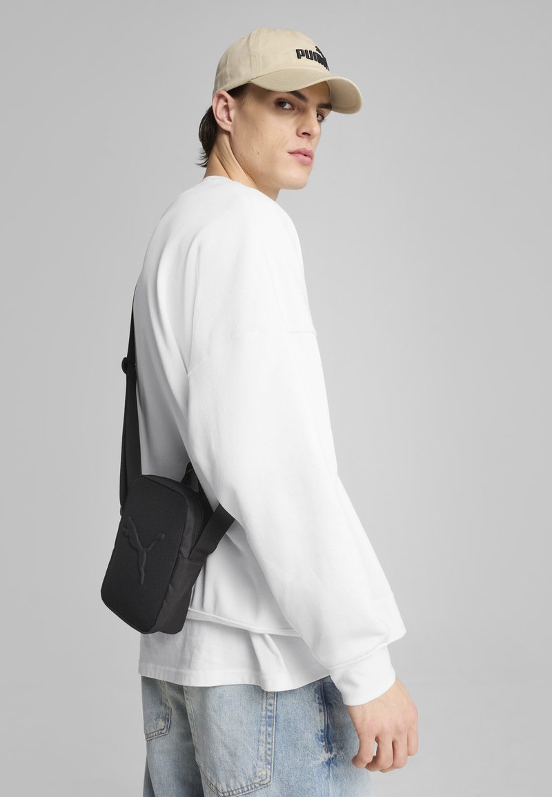 Sac à bandoulière noir avec logo, porté sur un sweat-shirt blanc et un jean bleu clair. Il présente un design rectangulaire élégant et une sangle réglable.