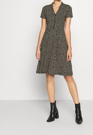 Woman wearing a black short-sleeve knee-length dress with white floral print and black heeled ankle boots standing against a plain background.