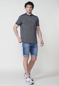 Jūras spēku un pelēks rakstains polo krekls ar pogājamu apkakli, pārī ar ruļļiem ievelcamiem džinsu šortiem un pelēkiem sporta apaviem ar tumšiem akcentiem.