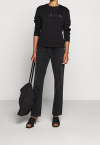 Schwarzer Sweatshirt mit geprägtem "HUGO"-Logo, kombiniert mit schwarzen Jeans mit geradem Bein. Das Model trägt eine schwarze Tote Bag und schwarze offene High Heels.