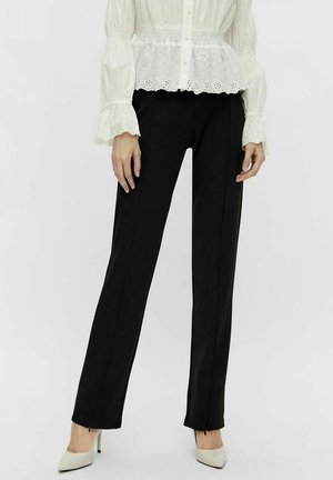 Kobieta ubrana w czarne spodnie o prostych nogawkach, zestawione z białą, długorękawową bluzką i białymi szpilkami z ostrym noskiem.