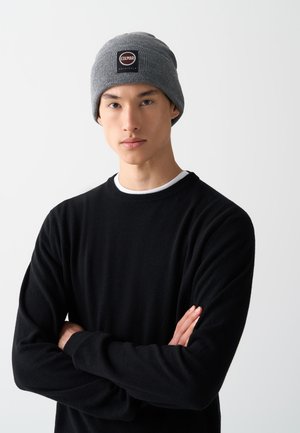 Căciulă tricotată gri cu o etichetă logo, purtată de un model într-un pulover negru. Materialul pare moale cu o textură înțepată.