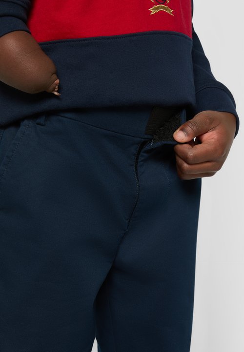 Pantalones azul marino con una textura suave y cierre de cremallera, con una lengüeta de velcro negra en la cintura. Suéter rojo y azul marino visible arriba.