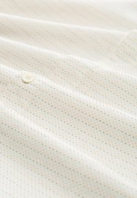 Weißer Stoff mit einem sich wiederholenden Muster aus kleinen schwarzen und orangen Punkten; im Vordergrund befindet sich ein weißer Knopf, weiche Textur.