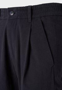 Dark blue cotton trousers with pleats, featuring a stitched waistband and a single button closure. Smooth texture with deep side pockets.