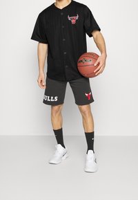 Jersey de béisbol de manga corta a rayas negras con botones, con un logo rojo. Pantalones cortos grises con el texto y el logo de "HULLS". Zapatillas blancas.