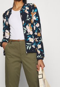 Kvinna klädd i olivgröna byxor med brunt bälte, vit tajt topp, marinblå blommig zip-up jacka, håller en krämfärgad texturerad handväska.