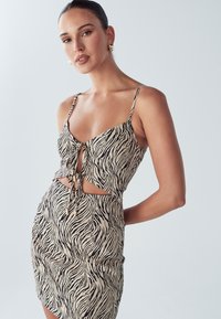 Zebramuster Kleid in Beige und Schwarz, mit einer Bindung vorne, ausgeschnittenen Seiten, dünnen Trägern und einer figurbetonten Silhouette in Mini-Länge.