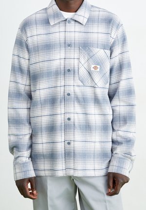 Homme portant une chemise à carreaux bleu clair et blanche à boutons, avec poche poitrine et petit logo de la marque, associée à un pantalon gris clair.