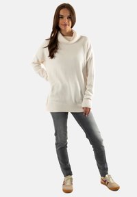 Suéter de punto blanco, de corte grande y con cuello alto, combinado con jeans ajustados grises y zapatillas beigas con acentos en color burdeos.