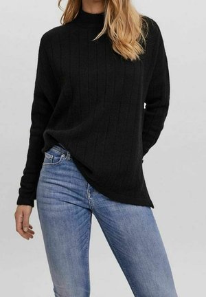 Femme portant un pull noir côtelé à col haut et un jean bleu clair ajusté, debout devant un fond uni clair.