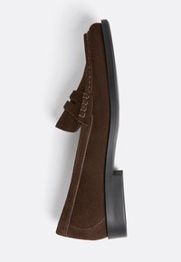 Braune Wildlederschuhe mit einer niedrigen, flachen schwarzen Sohle und einem dekorativen Riemen über der Oberseite. Seitennähte und klare Linien zeichnen das Design aus.