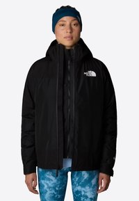 Kobieta nosząca czarną kurtkę North Face z kapturem, niebieskie leginsy w wzory oraz granatową opaskę na głowę, stojąca na jednolitym tle.