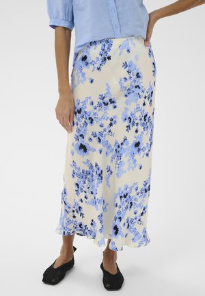 Žena v krémovej midi sukni s modrým kvetinovým vzorom, svetlomodrej blúzke s nafúknutými rukávmi a čiernych tkaných balerínkach.