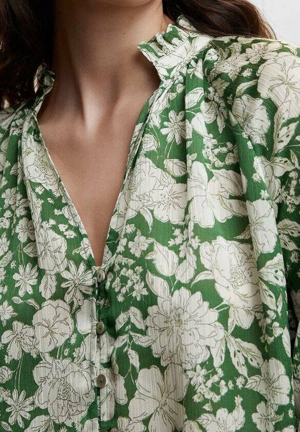 Femme portant un chemisier vert avec des motifs floraux blancs, comportant un col à volants et des boutons sur le devant.