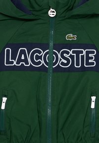 Veste légère verte avec fermeture éclair, panneau marine affichant "LACOSTE" et logo crocodile brodé. Dispose de deux poches zippées blanches.