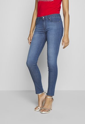 Vaqueros ajustados azules llevados por una persona de pie, combinados con sandalias plateadas de tacón alto y un top rojo con diseño blanco.