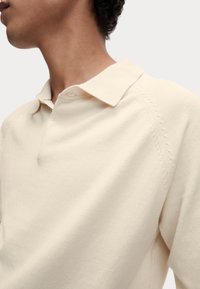 Nahaufnahme einer Person, die ein cremefarbenes, kragenloses Kurzarmhemd trägt, mit sichtbarer Stoffstruktur und Nähdetails an der Schulter.