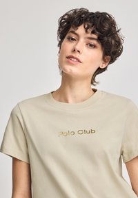 Beige t-shirt med kort ärm, tillverkad av bomull, med en guldfärgad "Polo Club"-logotyp i mitten. Rund halsringning och textil med struktur.