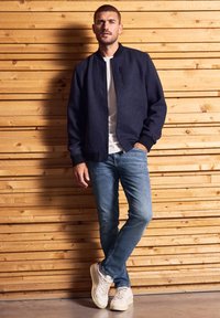 Hombre de pie contra una pared de panel de madera, vestido con una chaqueta azul marino, una camiseta blanca, jeans azules y zapatillas blancas, con una mano en el bolsillo.