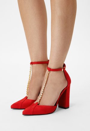 Røde ruskinds-højhælede sko med spids tå, ankelstropper og guldkæde som pynt. Tykkelse hæl for stabilitet.