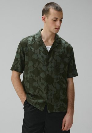 Mladý muž s krátkymi vlasmi, oblečený v zelenej kvetinovej košeli s krátkym rukávom, cez bielu podkošelu, stojaci s jednou rukou v vrecku.