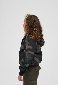 Veste camouflage dans des nuances de noir et de vert, avec une capuche et des poignets en tricot, vue de côté, portée avec un pantalon vert olive.