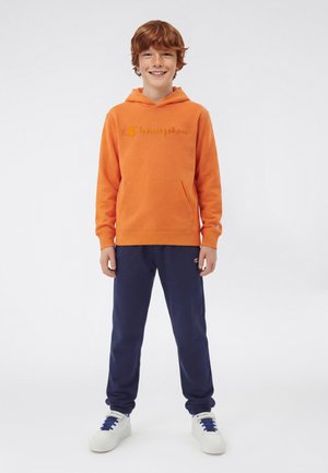 Niño sonriente con cabello rojo que lleva una sudadera con capucha Champion naranja, pantalones deportivos azul marino y zapatillas blancas, de pie contra un fondo liso.