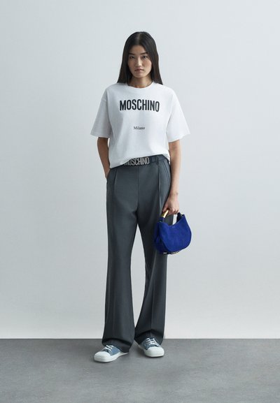 Naine kannab valget Moschino t-särki, tumehalli laia lõikega pükse, siniseid tossusid ja hoiab käes väikest sinist käekotti kuldsete käepidemetega.