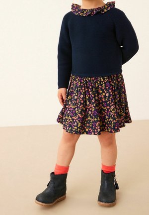 Pull en maille bleu marine avec col à volants, associé à une jupe patineuse à motif floral dans des tons bleu foncé, rose et jaune. Bottines noires et chaussettes orange.
