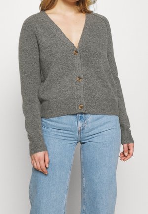Vrouw draagt een grijze, met knopen gesloten V-hals cardigan en lichtblauwe, hoog getailleerde jeans, staand tegen een witte achtergrond.