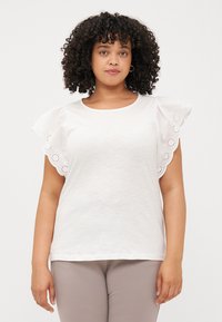 A woman with curly hair wears a white blouse with ruffled sleeves and lace detailing, standing in front of a neutral background.