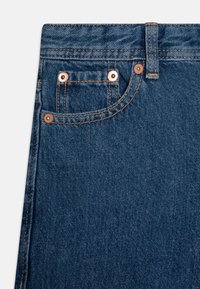 Temno modra denim tkanina z visoko pasom, bakrenimi nitmi in oranžnim strojenjem. Vključuje majhen sprednji žep in zanke za pas.